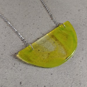Könnte beinhalten: Halskette mit limettengrünem Anhänger und silberner Kette. Der Anhänger hat eine halbkreisförmige Form mit transluzentem, marmoriertem Aussehen. Die Halskette wird auf einer hellgrauen Oberfläche präsentiert.
