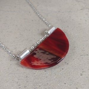 Könnte beinhalten: Eine Halskette mit einem halbmondförmigen Anhänger in Rot- und Brauntönen, der an einer feinen Silberkette befestigt ist. Der Anhänger hat eine glänzende Oberfläche und ist mit silbernen zylindrischen Verbindern an der Kette befestigt. Die Halskette liegt auf einer hellgrauen Oberfläche.