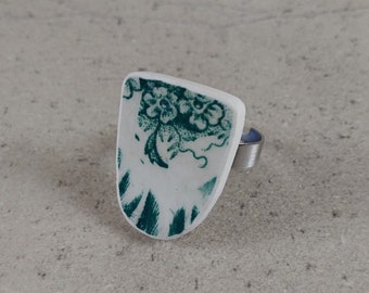 May include: A white ring with a green floral design on the top. The ring is made from a piece of broken china.