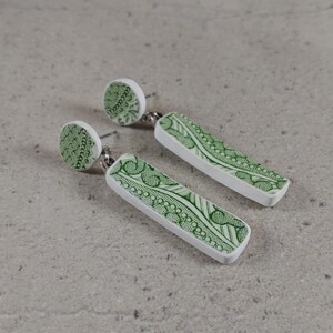 Peut inclure: Une paire de boucles d'oreilles pendantes avec une base blanche et un motif vert. Les boucles d'oreilles ont un petit sommet rond et un fond rectangulaire plus long.