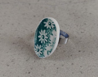 May include: A white oval ring with a green floral design. The ring is made of metal and has a silver band.