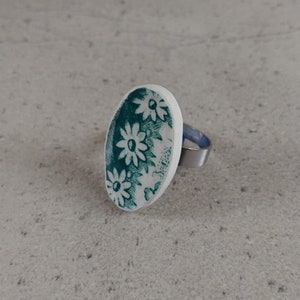 Könnte beinhalten: Ein weißer ovaler Ring mit grünem Blumendesign. Der Ring ist aus Metall und hat ein silbernes Band.