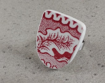 May include: A white and red patterned ring with a silver band. The ring features a rectangular design with a red floral pattern on a white background. The ring is set against a gray background.