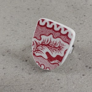 Könnte beinhalten: Ein Ring mit weißem und rotem Muster und einem silbernen Band. Der Ring hat ein rechteckiges Design mit einem roten Blumenmuster auf weißem Grund. Der Ring ist vor einem grauen Hintergrund platziert.