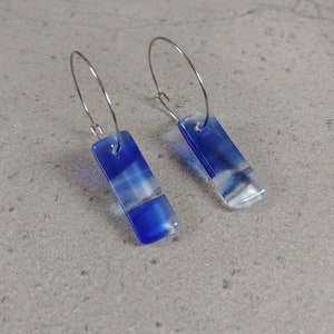 Peut inclure: Une paire de boucles d'oreilles créoles argentées avec des breloques rectangulaires en verre bleu et blanc. Les breloques ont un effet nuageux et marbré.
