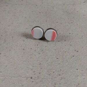 Puede incluir: Dos pendientes redondos con un diseño degradado blanco y rosa. Los pendientes tienen un borde de metal plateado. Los pendientes se muestran sobre una superficie gris.