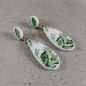 Peut inclure: Une paire de boucles d'oreilles en forme de goutte d'eau blanches avec un motif floral vert. Les boucles d'oreilles ont un petit dessus rond blanc avec un motif floral vert.