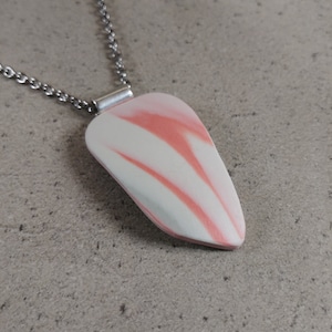 以下が含まれることがあります： 涙の形をしたペンダントが付いたネックレス。ペンダントは、白いベースにピンクの渦巻き模様が入っています。銀色のチェーンに吊り下げられています。ネックレスは、質感のあるニュートラルな色の表面に表示されています。