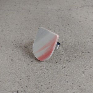 Könnte beinhalten: Ein Ring mit einem abstrakten Design in Weiß und Rosa. Der Ring ist aus Metall und hat eine glatte, abgerundete Form.