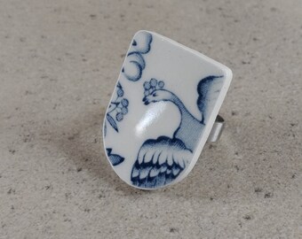 May include: A white ceramic ring with a blue and white floral design featuring a bird. The ring is set on a silver band.