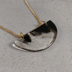 Peut inclure: Un collier en chaîne dorée avec un pendentif en résine transparente. Le pendentif a la forme d'une demi-lune et contient des tourbillons noirs et blancs.