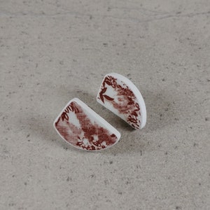 Puede incluir: Un par de pendientes de cerámica blanca con un diseño abstracto rojo y blanco. Los pendientes tienen forma de semicírculos.
