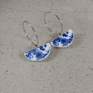 Peut inclure: Une paire de boucles d'oreilles créoles argentées avec des pendentifs en porcelaine bleu et blanc en forme de demi-cercle ornés d'un motif floral.