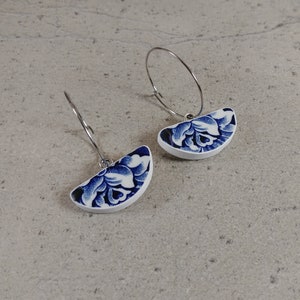 Peut inclure: Une paire de boucles d'oreilles en porcelaine bleue et blanche avec un motif floral. Les boucles d'oreilles sont en forme de demi-cercles et sont attachées à des anneaux argentés.
