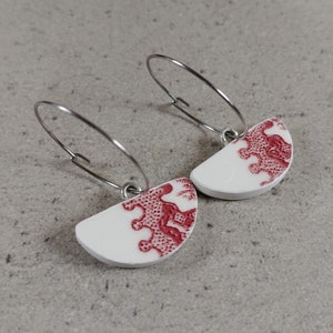 Peut inclure: Une paire de boucles d'oreilles blanches et rouges. Les boucles d'oreilles sont en forme de demi-cercle avec un motif floral rouge sur fond blanc. Elles sont suspendues à des fermoirs à créoles argentés. Le fond est une surface gris clair.