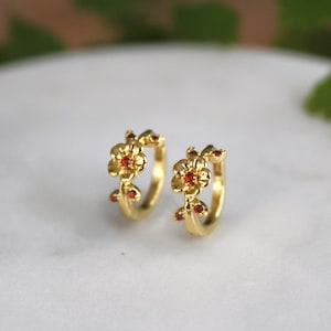 May include: A pair of gold-colored hoop earrings. Each earring features a floral design with red gemstone accents. The earrings are set against a white marble surface with green foliage in the background.