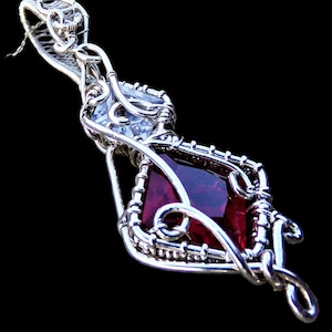 May include: A silver wire wrapped pendant featuring a red gemstone and a clear crystal. The pendant is designed in a geometric shape with intricate wirework.