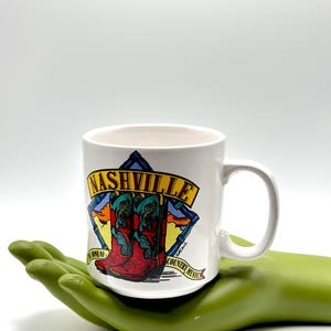 May include: White ceramic mug featuring a colorful illustration of a cowboy boot with the word "NASHVILLE" in yellow. The mug also has the text "HOME OF COUNTRY MUSIC". The mug is held by a green hand-shaped object.