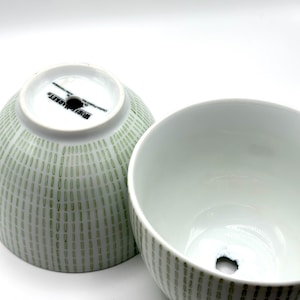 May include: Two white ceramic bowls with a green dotted pattern. One bowl is upright, and the other is upside down, revealing a hole in the center. The bowls have a smooth, glossy finish and are set against a white background.