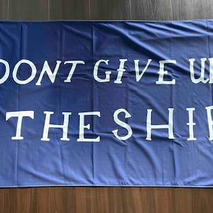 Può includere: Una bandiera blu navy con testo bianco che recita "DON'T GIVE UP THE SHIP". La bandiera ha occhielli agli angoli per appenderla. La bandiera è rettangolare e sembra essere fatta di un tessuto leggero.