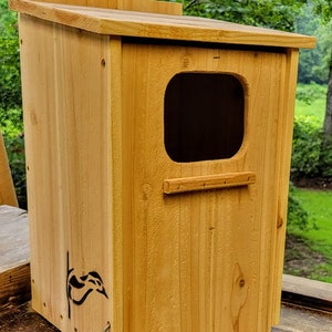 May include: A wooden birdhouse with a small opening and a perch. The birdhouse is painted a light brown color and has a small, black logo on the front.
