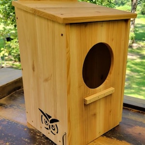 May include: A wooden birdhouse with a sloped roof and a large circular entrance. The birdhouse is made of light-colored wood and features a decorative owl silhouette. A small wooden perch is below the entrance.