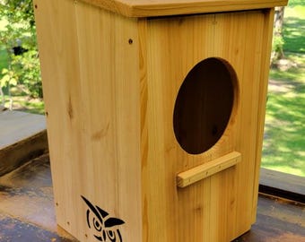 May include: A wooden birdhouse with a sloped roof and a large circular entrance. The birdhouse is made of light-colored wood and features a decorative owl silhouette. A small wooden perch is below the entrance.