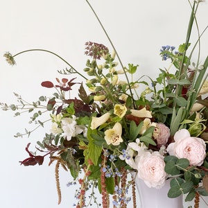 Pode incluir: Um arranjo floral exuberante em uma urna branca. O buqu&ecirc; apresenta rosas rosa claro, flores brancas e amarelas e v&aacute;rias folhagens. O arranjo &eacute; colocado em um fundo branco.