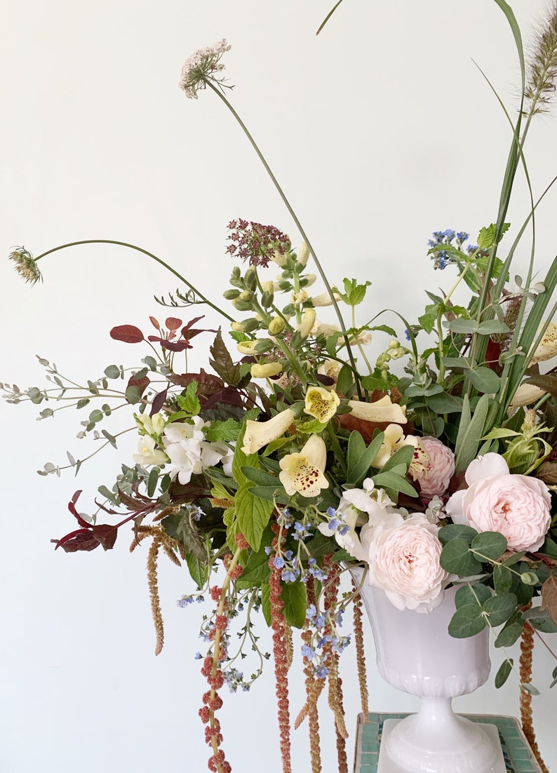 Pode incluir: Um arranjo floral exuberante em uma urna branca. O buqu&ecirc; apresenta rosas rosa claro, flores brancas e amarelas e v&aacute;rias folhagens. O arranjo &eacute; colocado em um fundo branco.