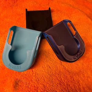 May include: Three 3D-printed holders. One is light blue, another is dark blue, and the third is black. The holders have a curved shape with a handle and a slot. The holders are on an orange surface.