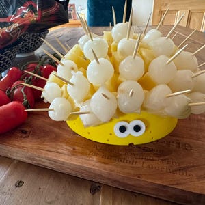 May include: A creative food presentation resembling a hedgehog. It features pineapple chunks, white melon balls, and skewers, presented on a yellow plate with googly eyes. Red tomatoes are on a wooden board.