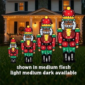 May include: Three illuminated nutcracker figures in different sizes, standing on a green lawn. The largest nutcracker is red and green with a black face and white beard. The middle nutcracker is red and green with a black face and white beard. The smallest nutcracker is red and green with a black face and white beard. The text "shown in medium flesh light medium dark available" is at the bottom of the image.
