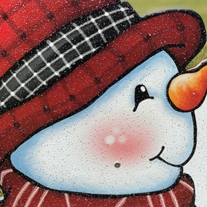 a Christmas snowman lawn sign: A close-up of a snowman's face. The snowman has a red hat with a black and white plaid band, a red scarf, and a white face with rosy cheeks. The snowman's nose is orange and the eyes are black. The snowman's mouth is slightly open and the face is covered in white glitter.