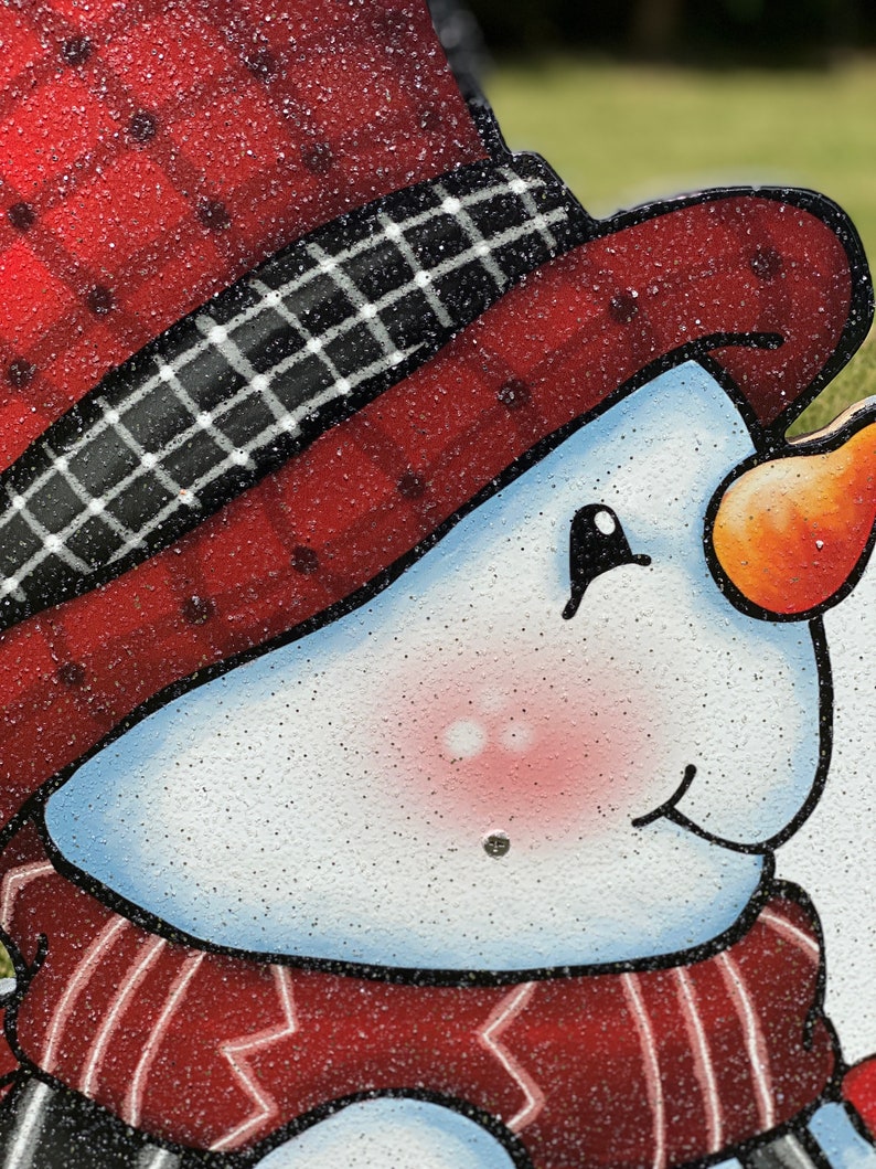 a Christmas snowman lawn sign: A close-up of a snowman's face. The snowman has a red hat with a black and white plaid band, a red scarf, and a white face with rosy cheeks. The snowman's nose is orange and the eyes are black. The snowman's mouth is slightly open and the face is covered in white glitter.