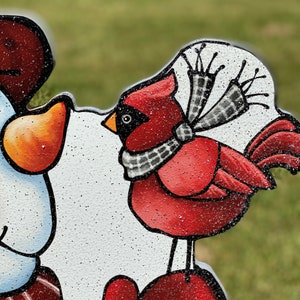 a Christmas snowman lawn sign: close up view of the red cardinal wearing a black and white scarf is perched on a red mitten. The cardinal is painted on a white background with glitter.