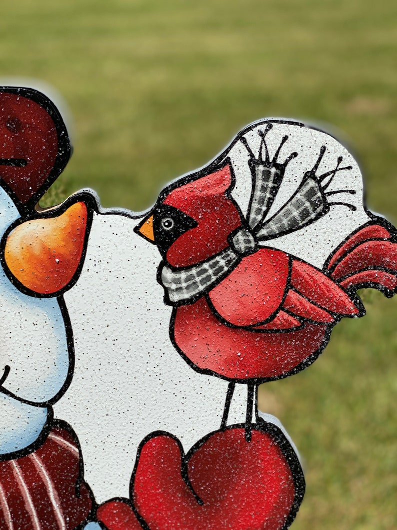 a Christmas snowman lawn sign: close up view of the red cardinal wearing a black and white scarf is perched on a red mitten. The cardinal is painted on a white background with glitter.
