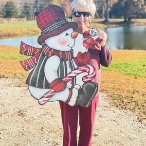 an adorable snowman yard sign, complete with a candy cane, cozy scarf, and a sweet cardinal perched on his mitten: A snowman wearing a red and black plaid hat, a red scarf, and a black and white striped vest. The snowman is holding a red and white candy cane and is looking at a red cardinal with a white ribbon around its neck.