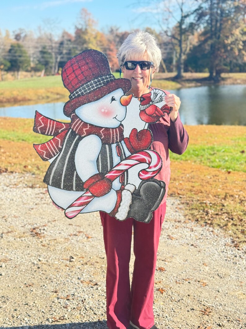 an adorable snowman yard sign, complete with a candy cane, cozy scarf, and a sweet cardinal perched on his mitten: A snowman wearing a red and black plaid hat, a red scarf, and a black and white striped vest. The snowman is holding a red and white candy cane and is looking at a red cardinal with a white ribbon around its neck.