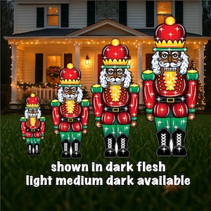 May include: Three illuminated nutcracker figures in various sizes, standing on a green lawn in front of a house. The nutcrackers are red, green, and black with white accents. The largest nutcracker is in the center, and the smallest is on the left. The text "shown in dark flesh light medium dark available" is at the bottom of the image.
