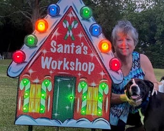 May include: A triangular Santa's Workshop sign featuring red, green, and white colors with candy cane graphics, a central door, and illuminated multi-colored bulbs along the edges. The sign is mounted on two green metal stakes for outdoor display.