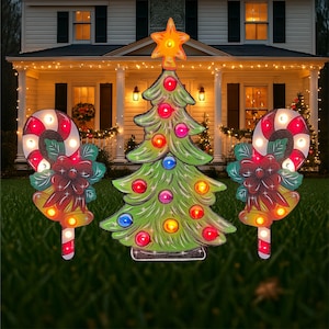 May include: A set of three lighted Christmas yard decorations featuring a green Christmas tree, a red and white candy cane, and a red poinsettia with a gold bell. The decorations are illuminated with colorful lights.