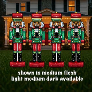 May include: Three illuminated nutcracker figures stand on red bases in a grassy yard. The nutcrackers are wearing green jackets, red pants, and black boots. The figures are lit with white lights. The text "shown in medium flesh light medium dark available" is at the bottom of the image.