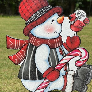 an adorable snowman yard sign, complete with a candy cane, cozy scarf, and a sweet cardinal perched on his mitten: A snowman wearing a red and black plaid hat, a red scarf, and a black and white striped vest. The snowman is holding a red and white candy cane and is looking at a red cardinal with a white ribbon around its neck.