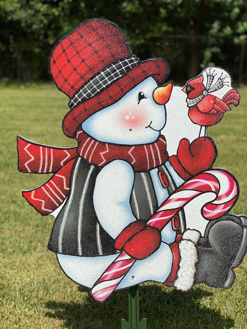 an adorable snowman yard sign, complete with a candy cane, cozy scarf, and a sweet cardinal perched on his mitten: A snowman wearing a red and black plaid hat, a red scarf, and a black and white striped vest. The snowman is holding a red and white candy cane and is looking at a red cardinal with a white ribbon around its neck.