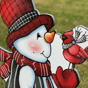 May include: A snowman with a red and black plaid hat, a red scarf, and red mittens holds a candy cane and a red cardinal.