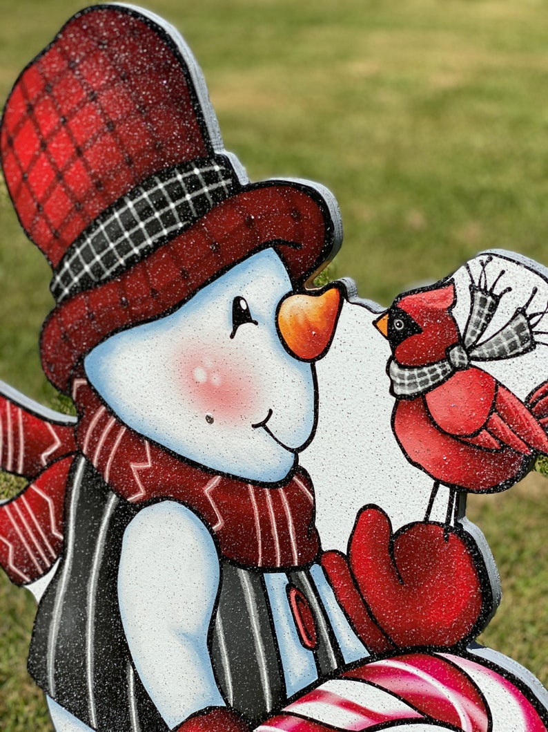 May include: A snowman with a red and black plaid hat, a red scarf, and red mittens holds a candy cane and a red cardinal.