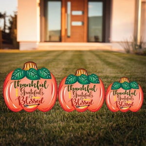 May include: Three orange pumpkin-shaped yard signs with the text "Thankful Grateful & Blessed" and an arrow pointing to the right. The signs are arranged in a row from largest to smallest.