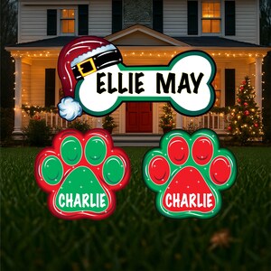 May include: A Christmas-themed yard decoration set with a bone-shaped sign that reads "Ellie May" and two paw print signs that read "Charlie". The bone sign has a Santa hat on top. The paw prints are red and green with glitter.