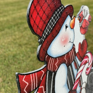 A Christmas snowman yard sign with a red and black plaid hat, a red and white scarf, and a red and white candy cane. The snowman is holding the candy cane in its right hand. The snowman is standing on a green lawn.