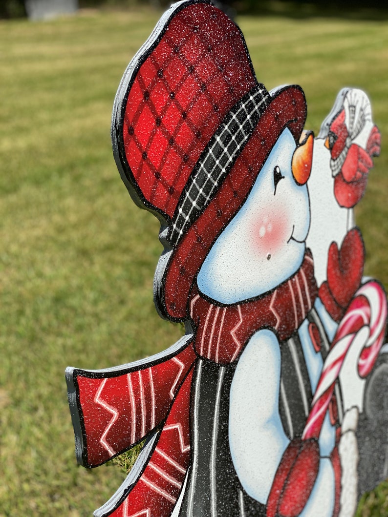 A Christmas snowman yard sign with a red and black plaid hat, a red and white scarf, and a red and white candy cane. The snowman is holding the candy cane in its right hand. The snowman is standing on a green lawn.